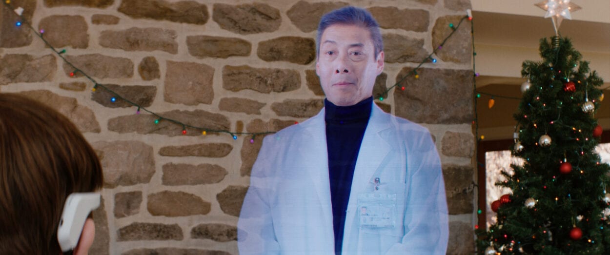 The Director (François Chau), a hologram, stands in front of a stone fireplace and Christmas decorations, addressing the couple.