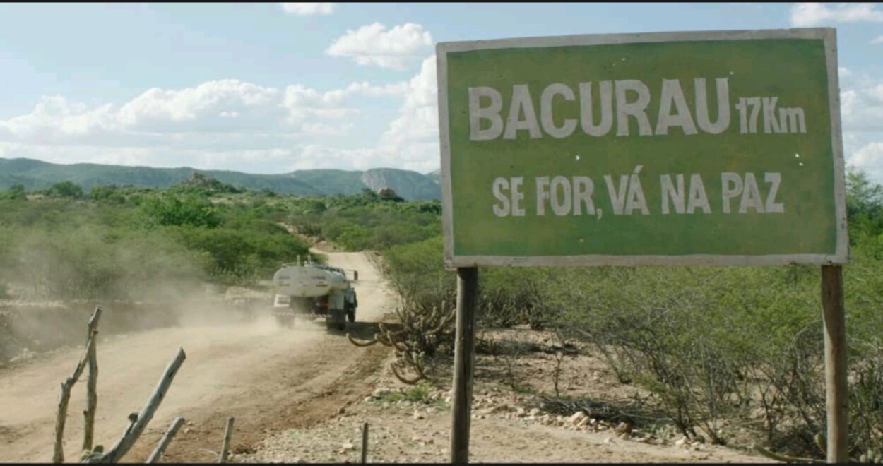 A medium shot showing a car driving past a signpost of Bacurau with a Portuguese sentence underneath the town's name stating "If you go, go in peace"