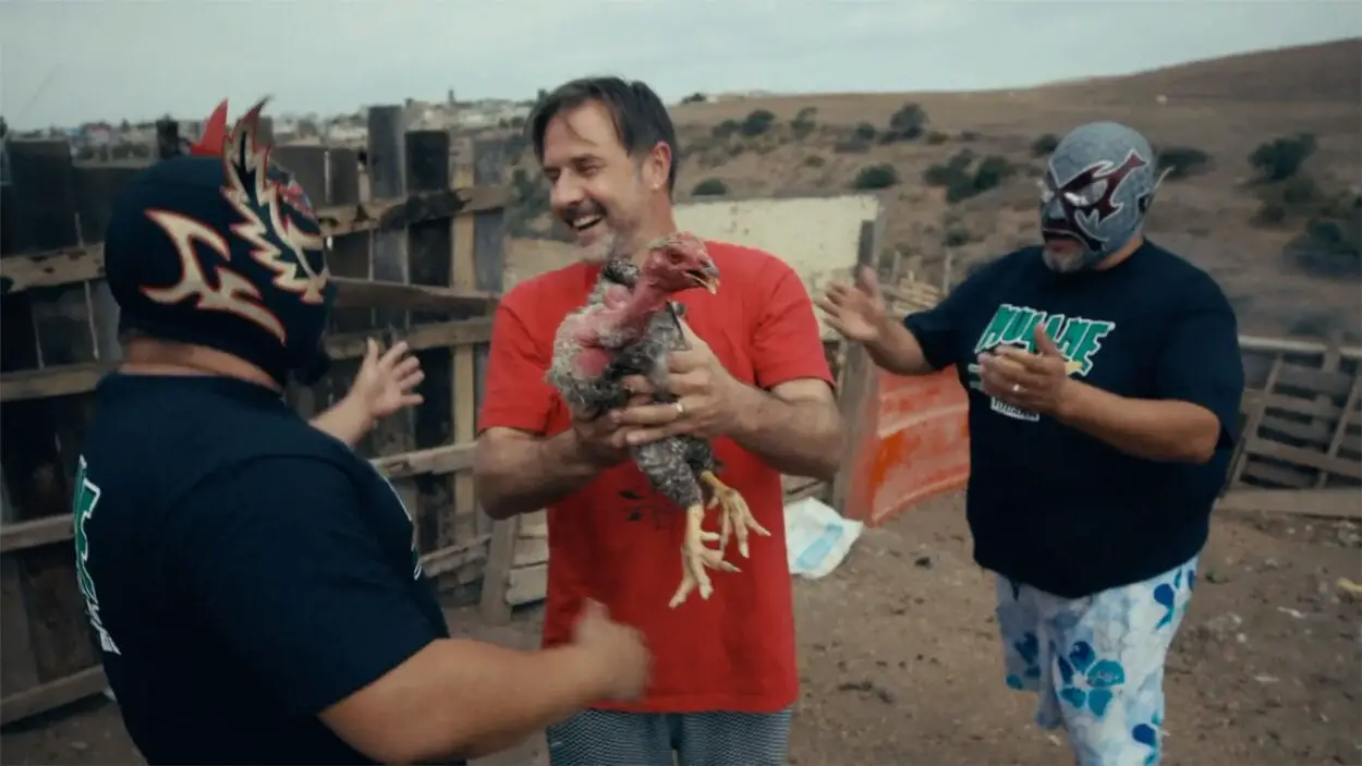 David Arquette standing in the Mexico desert, holding a chicken, surrounded by two luchadors.