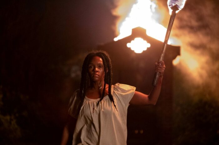 Eden stands with a torch in front of a shanty on fire in defiance.