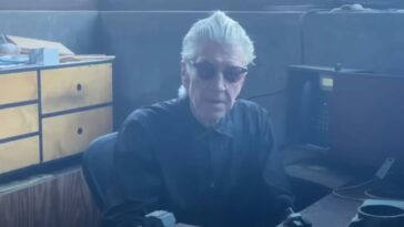 David Lynch sits at his desk wearing dark sunglasses