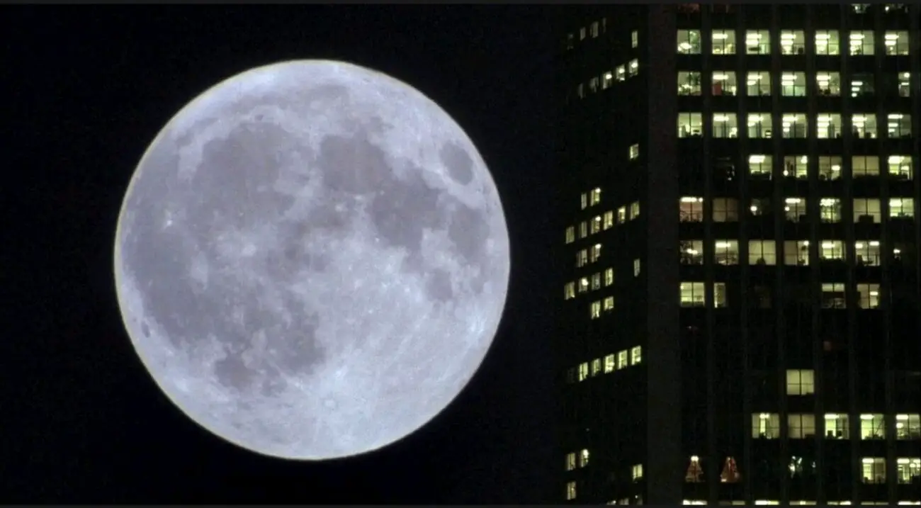 A long shot of the moon rising on the left side of an illuminated skyscraper.
