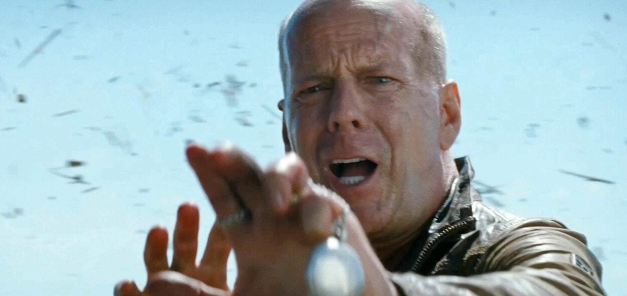 Bruce Willis holding his hands out with grass flying behind him