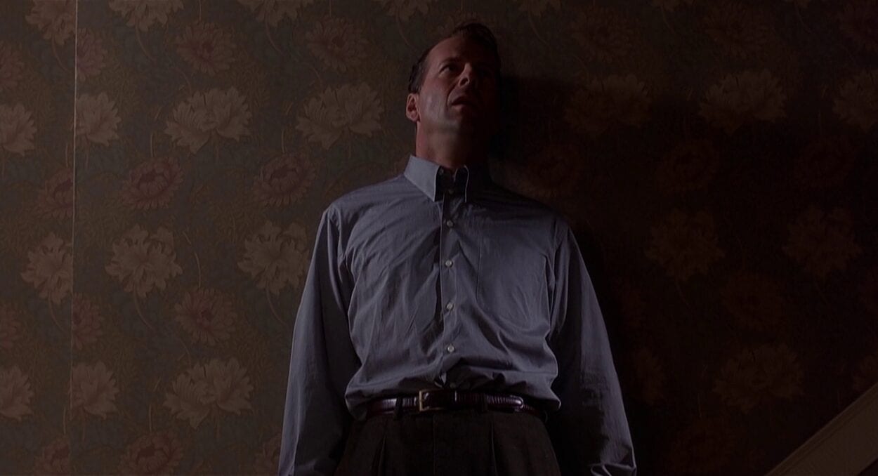 Bruce Willis standing atop the stairs, against the wall
