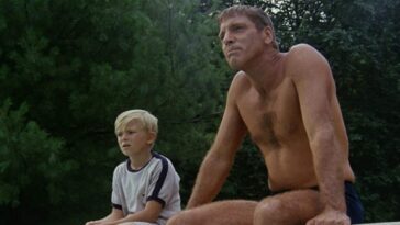 An image from The Swimmer 1968. Ned Merrill (Burt Lancaster) sits next to a young boy named Kevin Gilmartin Jr. on the edge of an empty swimming pool. Lancaster is shirtless and only wearing a small blue bathing suit. Both are staring into the distance.