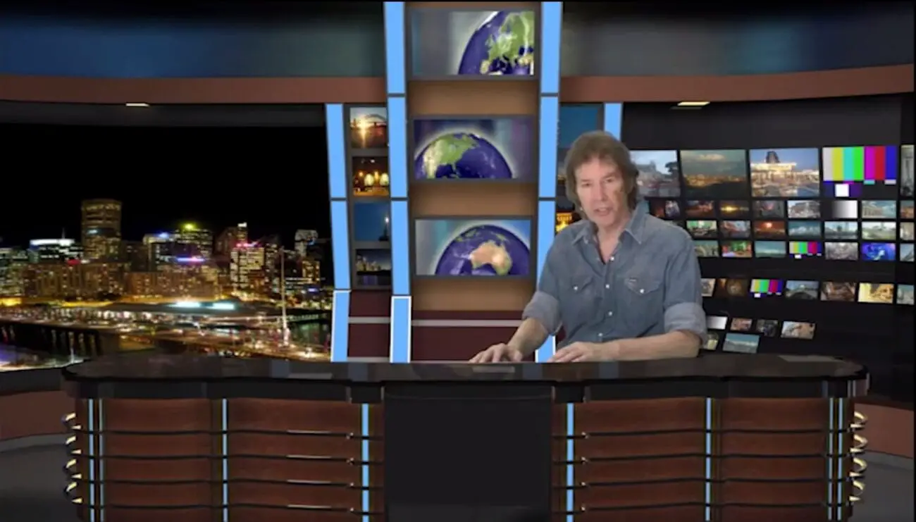 Neil Breen sits at a news desk to deliver his climactic speech at the end of Pass Thru. The background is an obvious green-screen.