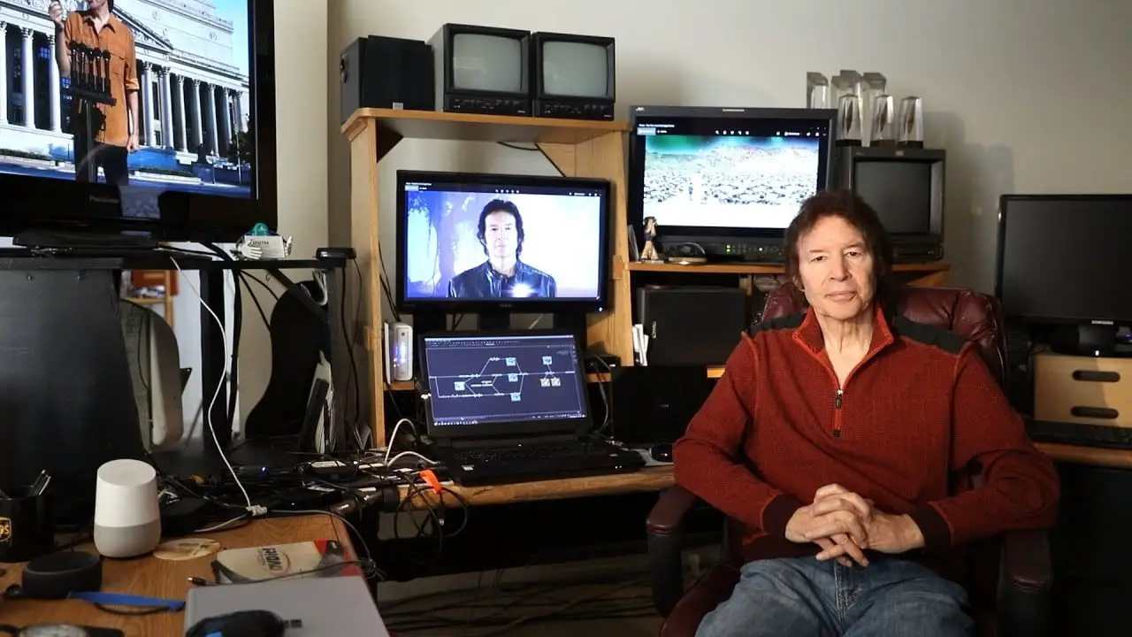 Neil Breen sits in his studio. Screens are on behind him with stills from his films.