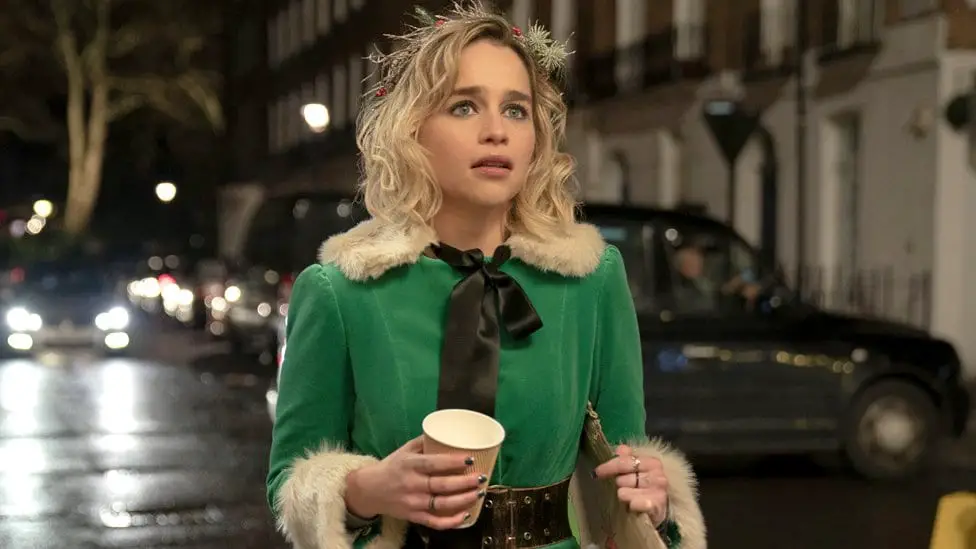 Emilia Clarke in her elf costume in Last Christmas, standing in the street with a cup of coffee, looking into the distance in dismay