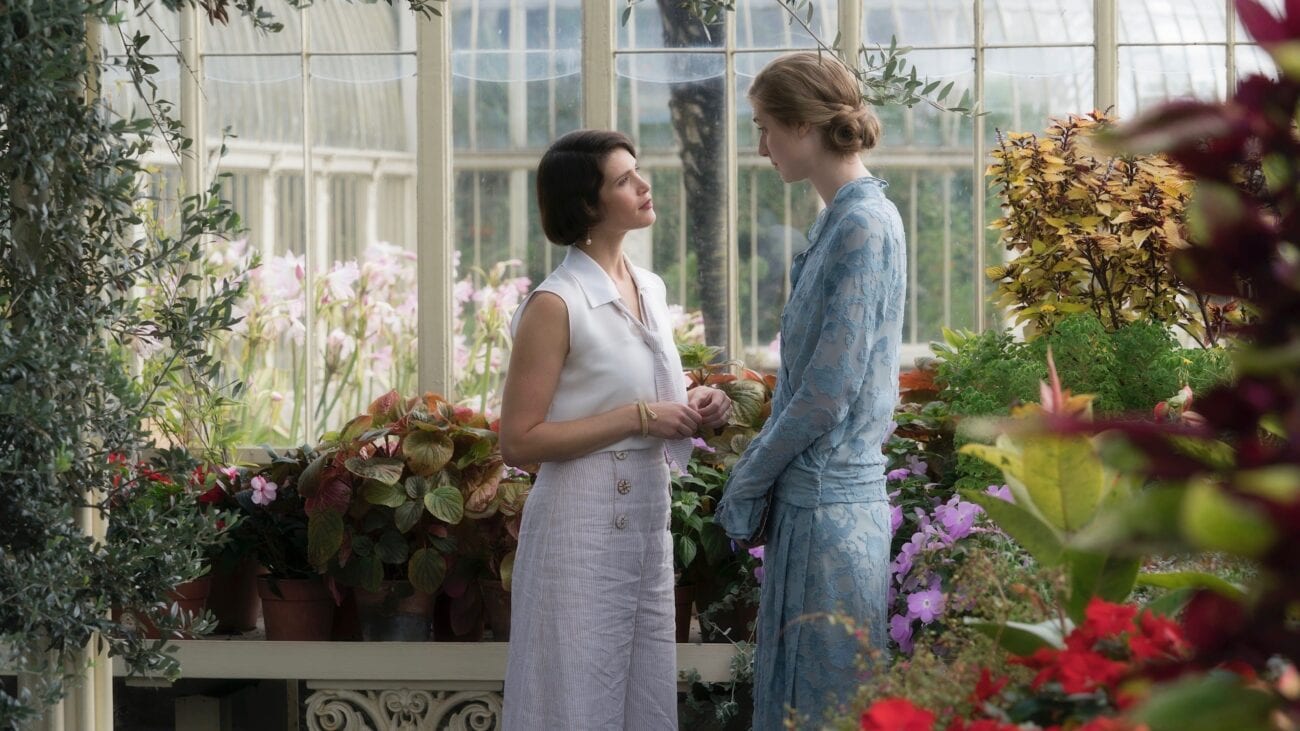 Vita and Virginia stand in a large glasshouse. They are surrounded by flowers and are looking at each other
