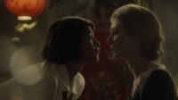 Vita and Virginia lean close to one another. The background is blurred but all the colours are warm