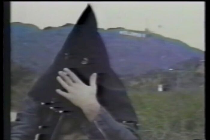 The hooded Eldon "El Duce" Hoke smokes a cigarette through his mask holes with the iconic Hollywood sign behind him