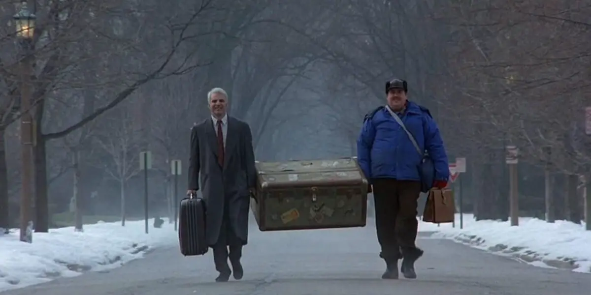 Neal and Del walking down a snowy street while carrying a trunk between them in Planes, Trains and Automobiles