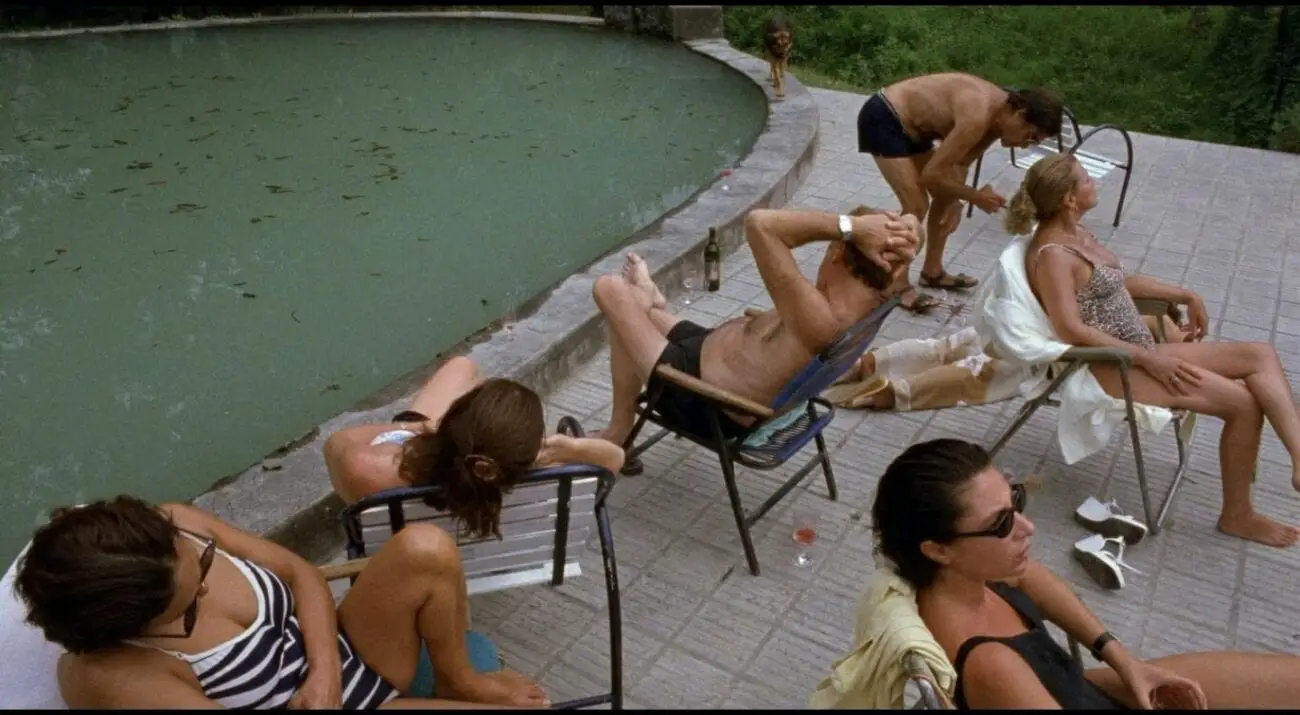 A group of adults sits by an unclean swimming pool with some facing towards the water, while other adults sit to their backs looking in the opposite direction.