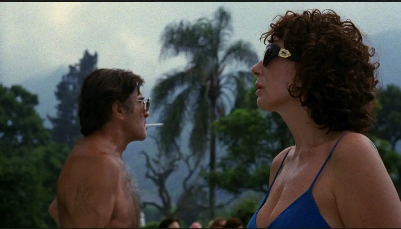 A medium shot of Gregorio (Martín Adjemián) smoking on the left of the frame while Mecha (Graciela Borges) stands on the right while they both look forward in their direction.