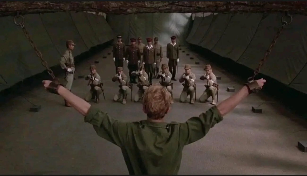 A wise shot behind Major Celliers (David Bowie) as his arms are raised by chains while a Japanese firing squad aims towards him