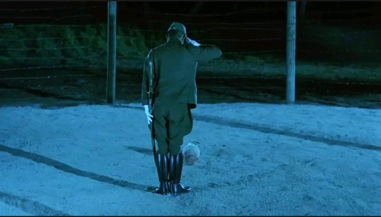 A long shot of Captain Yanoi (Ryuichi Sakamoto) as he stands and bows in front of the head of Major Celliers (David Bowie) as his body is buried underneath the sand