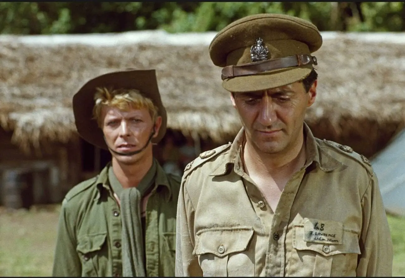 A medium shot of Colonel Lawrence (Tom Conti) looking down while Major Celliers (David Bowie) stands behind him and looks towards the camera