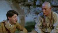 A medium shot of Colonel Lawrence (Tom Conti) sitting down beside Seargent Hara (Takeshi Kitano) while looking at him.