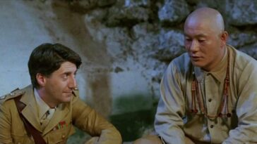 A medium shot of Colonel Lawrence (Tom Conti) sitting down beside Seargent Hara (Takeshi Kitano) while looking at him.