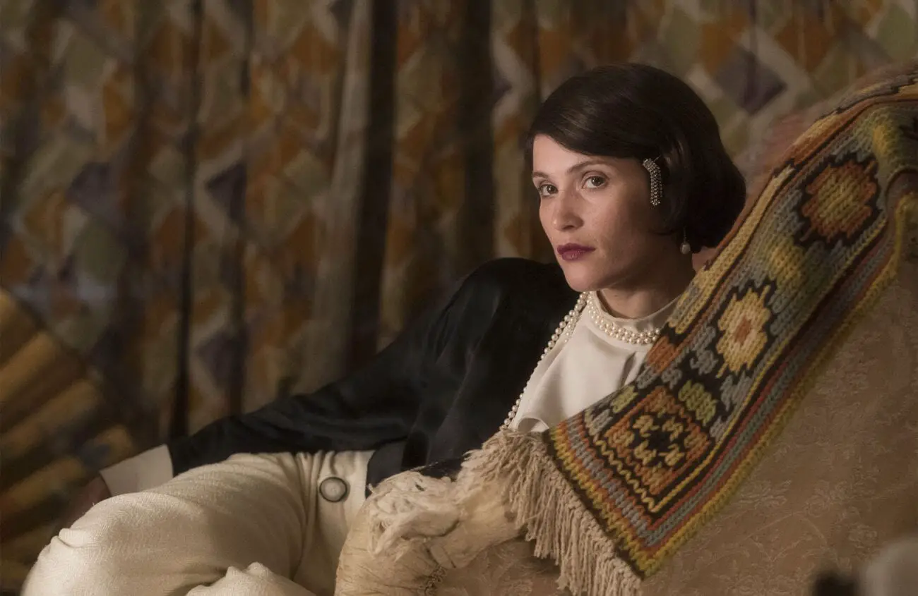 Gemma Arterton as Vita is wearing stylish clothing, including trousers, and is lounging on a seat