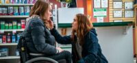 Diane squats down to comfort a wheelchair-bound Chloe in a pharmacy.