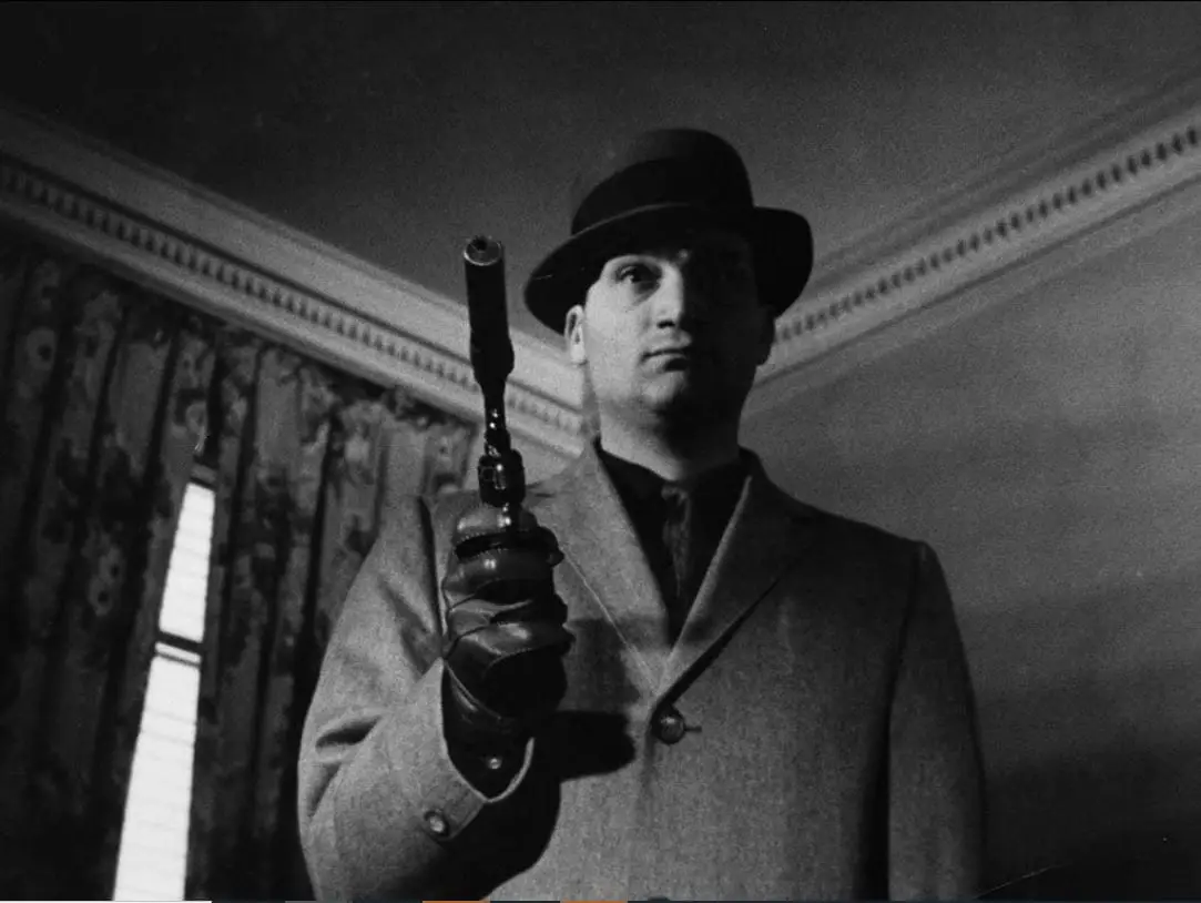 Still from Blast of Silence. Frankie Bono is shot from a low angel by the camera. He is wearing a fedora, trench coat, and holding a pistol with a silencer.