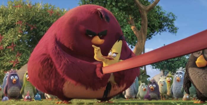 Terence (a very large bird, almost identical to Red but with darker coloring) preparing to launch Chuck (a small, vaguely triangle shaped yellow bird with long black tail and head feathers, a long orange beak and reddish brows) from a slingshot. Chuck is pointing a finger at him as though giving instructions. Terence looks angry.