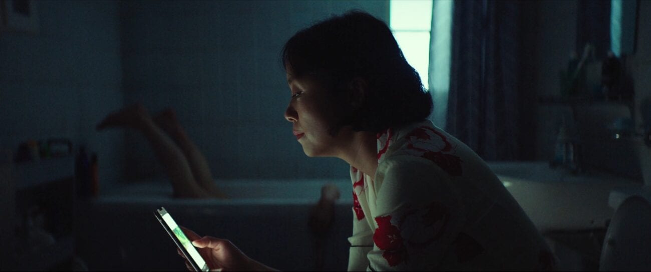 A woman in a red and white dress sits on a toilet looking at her phone in Beasts Clawing at Straws