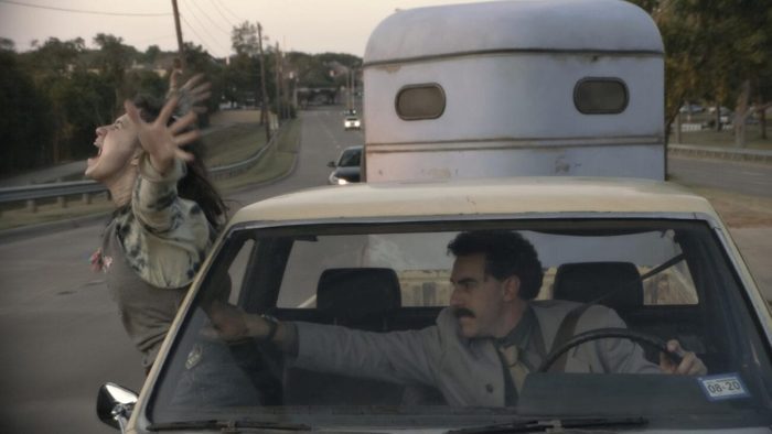 Borat, a man with curly black hair and a mustache, driving down a street, reaching to grab his daughter Tutar, a girl in dull-colored clothes with long dark brown hair, who is half out of the passenger window, her arms extended to either side of her while screaming.