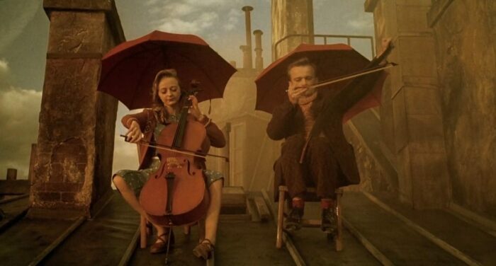 Julie and Louison sit on the rooftop playing instruments. Julie plays the cello while Louison plays a musical saw.