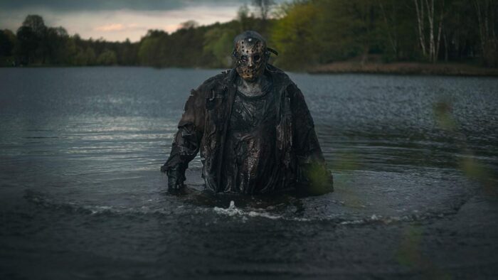 Jason Voorhees, hockey mask charred and clothes sodden, emerging from a lake at dawn. Whatever skin we can see is a deathly gray. There is some sort of blurry foliage in the bottom right corner, and the surrounding forest is also out of focus.