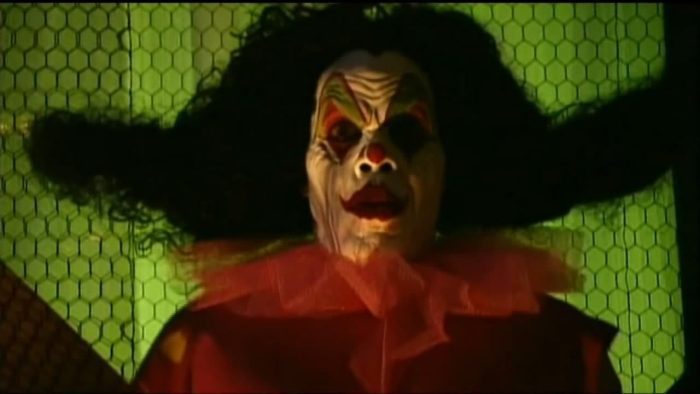 Killjoy, a wrinkly-faced demonic clown in white face paint with large red lips, a red dot on the end of his nose, and red and dramatic red and green eye makeup standing in front of a green background bearing a hexagonal design (looks to maybe be a fence). He has a cheap looking frilly red collar and a red suit. His hair is fuzzy and black, and on either side somewhat resembles two L's turned on their sides.