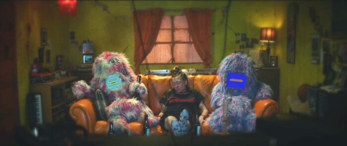 A girl with blonde hair, irradiated skin and pure white eyes sits on a couch between two colorful shaggy-furred beasts with screens for faces. They're in a dingy, yellow-lit living room with several bottles of beer and a pair of bongs.