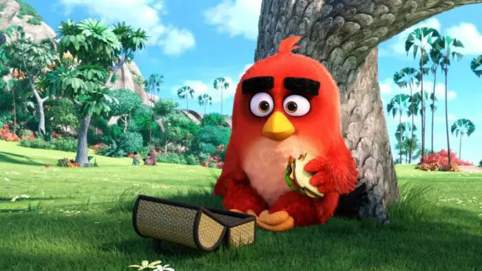 Angry Birds lead Red (a rather oval-shaped red bird with pinkish coloring on his chest/stomach, large rectangular black eyebrows, a yellow beak, fiery brown eyes, and orange feet) eating a sandwich under a tree. He's sitting on top of bright green grass, his wicker lunchbox in front of him. It's a bright and sunny day; a couple other birds can be seen sitting together in the background.
