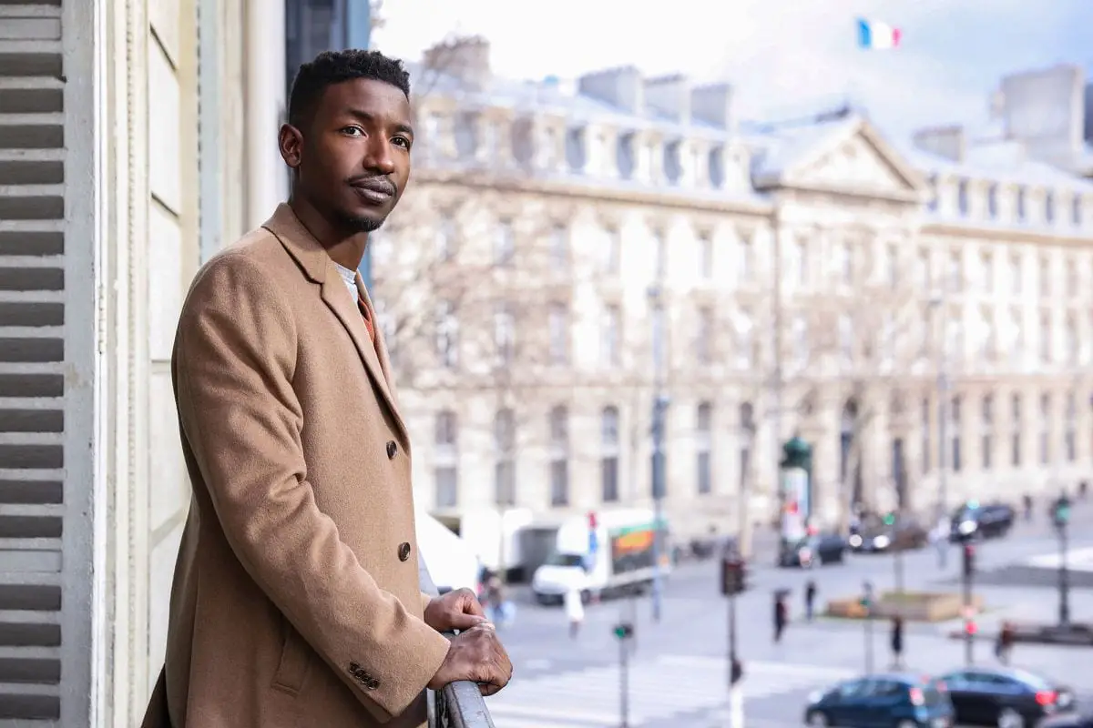 Elijah, a man with fuzzy black hair and thin facial hair, standing on a hotel balcony in Paris. He's wearing a light brown coat, hands resting on the balcony rails as he looks in the general direction of the camera. He looks happy.