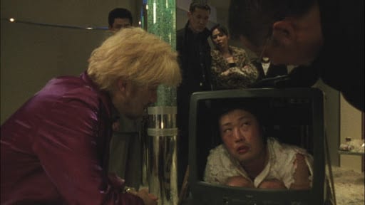 Kakihara (left), a man with shaggy blonde hair in a metallic purple coat, crouched to look at a black-haired man holed up inside a screenless VCR TV (middle). The TV man is looking to the right at another man in glasses and a black coat (right) bending down to look at him. Behind a pillar full of bubbly water are several other people looking at the scene.
