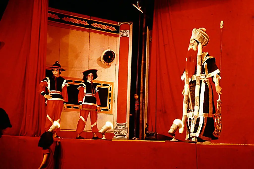 Three actors stand on stage in a Chinese Opera. The background and floor are red and the two off to the left are dressed as guards while the actor to the right is dressed in full ceremonial robes of the Emperor 