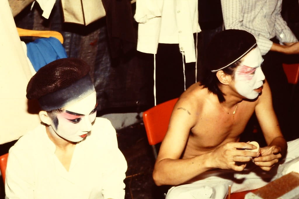 Backstage at a Chinese theatre, two performers are seen putting on traditional Chinese Opera make-up.