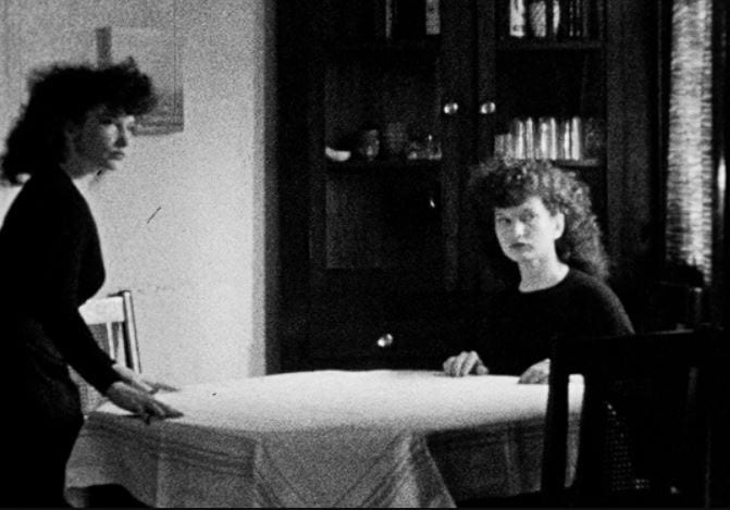 Two Maya Derens at a dinner table. One is standing as if about to sit down, the other is seated. They are both looking off-camera.