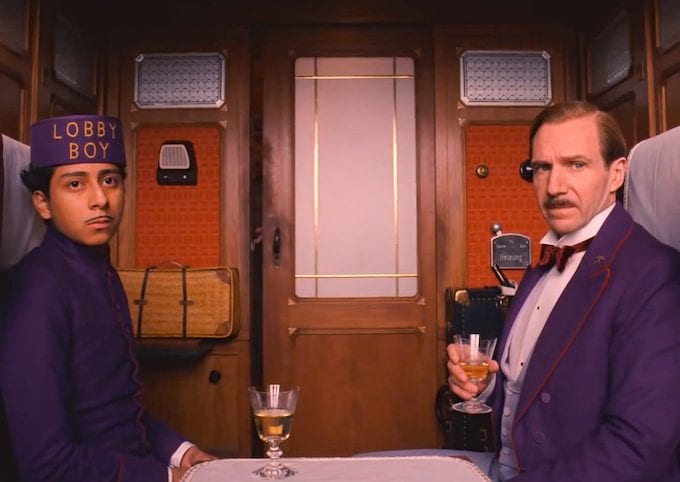 a young man wearing a purple uniform with "lobby boy" embroidered on the hat and a man in a purple tuxedo sit in a train car, looking at the camera. the man holds a small glass.