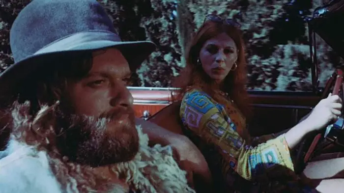 Mike and Elizabeth in American Hippie in Israel. Elizabeth is wearing a colorful shirt while driving. Mike is in the passenger seat wearing his wide-brimmed hat and goat vest.