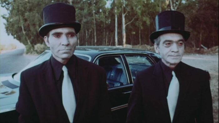 The two mystery men from American Hippie in Israel. Both are wearing black suits, white ties, top hats, and ghostly white face makeup.