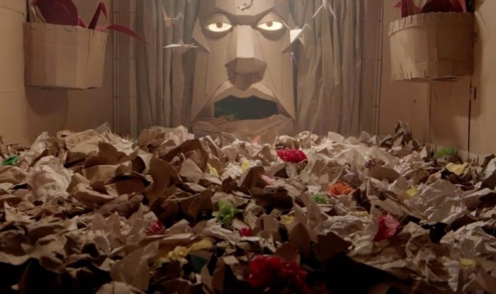 piles of cardboard and paper fill a room. in the back is a giant face set into the wall with glowing eyes, also made of cardboard