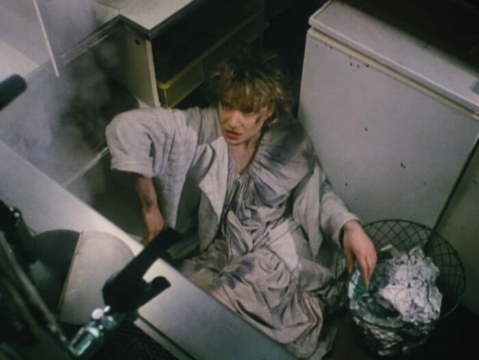 Fro above, woman cowers in a corner near a trash can filled with crumpled paper. She looks warily to some dissipating fog.