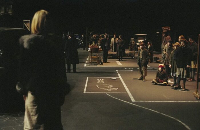 a woman walks on a empty stage, the ground marked with chalk. there are being sitting in the distance