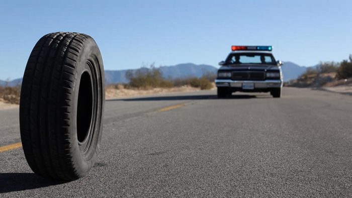 a tire rolls down a desert road followed by a police cruiser