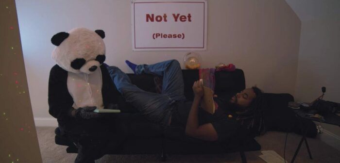 a person in a panda costume sits on a couch. Kamus is laying down writing in a notebook. behind them on the wall a poster says "not yet (please)"