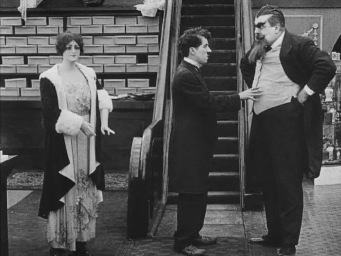 Still from The Floorwalker. Charlie Chaplin encounters Eric Campbell's general manager on the department store floor. They're standing in front of an escalator and mannequin.