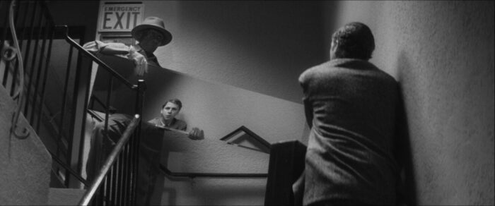 Criterion Channel Shorts March. Still from Man Rots from the Head. Michael Cera stands in front of a man moving a mirror in a stairwell. Cera's back is to us, but his face is reflected in the mirror.