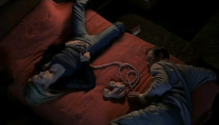 A medium overhead shot of Allegra Geller (Jennifer Jason Leigh) and Ted Pikul (Jude Law) laying together on a bed while connected to the umbilical cord shaped virtual reality technology